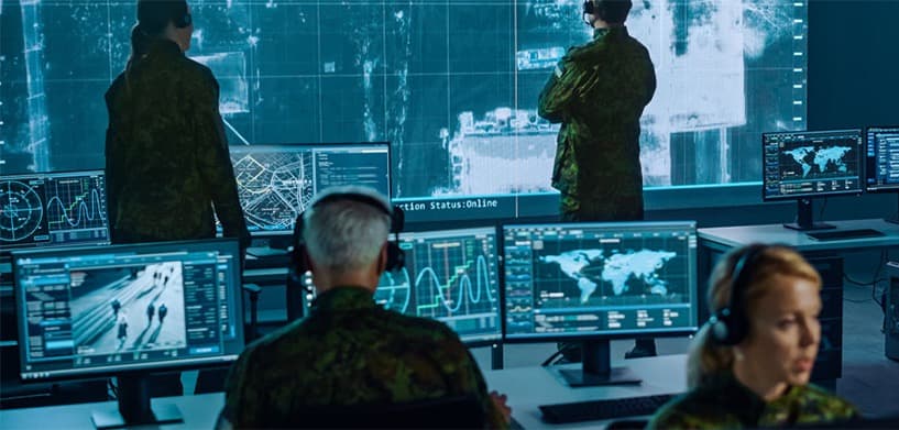 Military personnel monitoring a command-centre wall of live maps, radar feeds and analytics screens