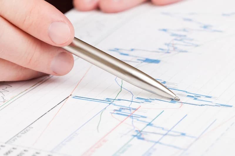 A hand holds a pen over a printed financial chart with rising and falling lines