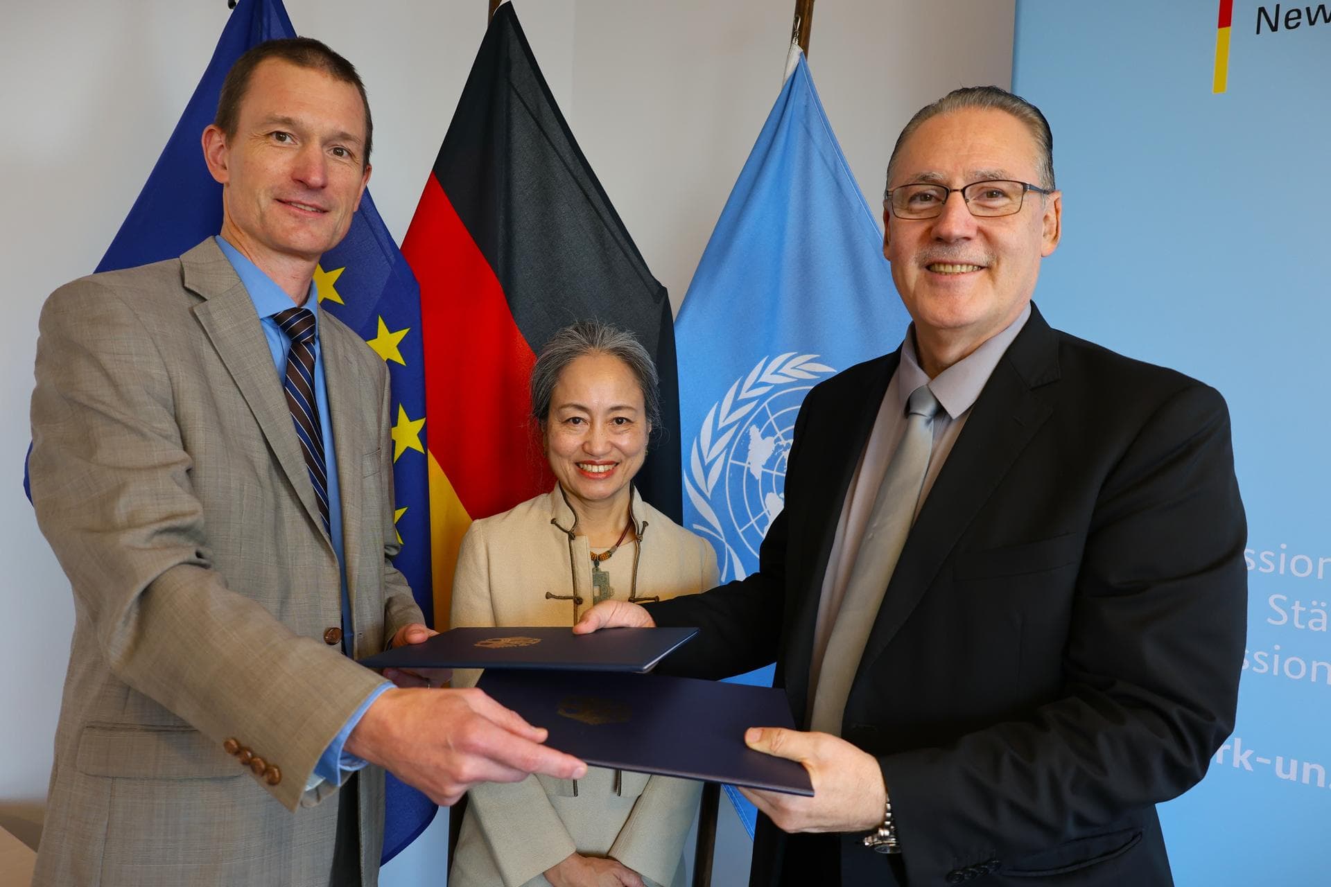 German and UN officials exchanging signed agreement folders in front of European Union, German, and United Nations flags