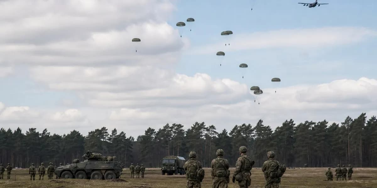 Paratroopers descending under parachutes over a German military training field as soldiers and armored vehicles watch from the ground