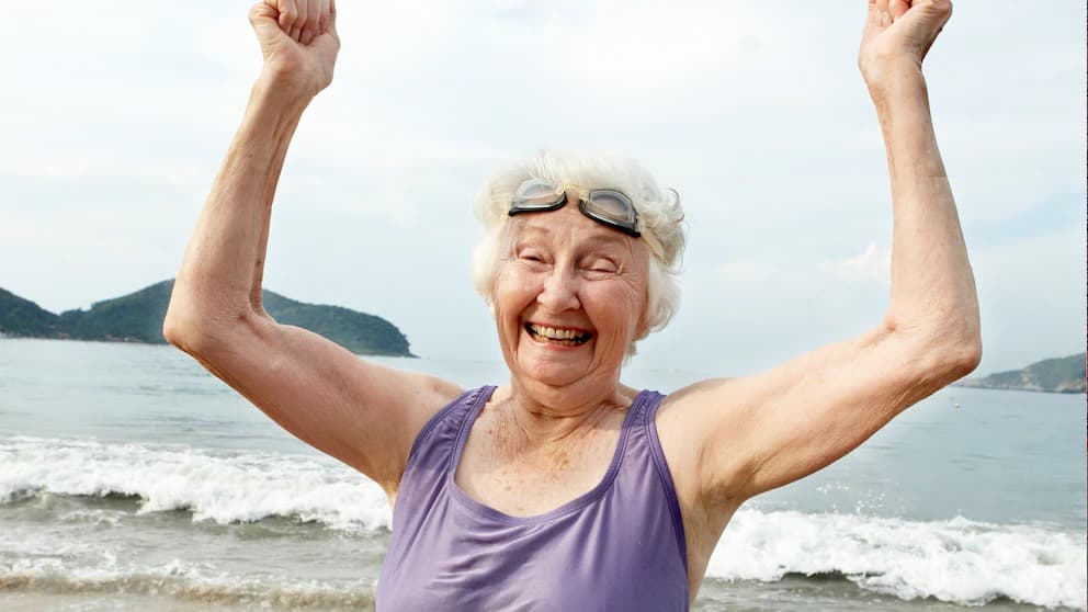 A smiling older woman raises her arms in triumph on a beach