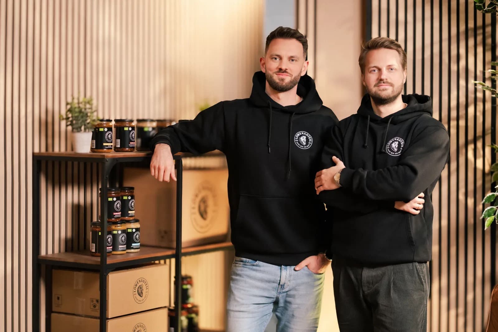 The two founders of Löwenanteil in branded hoodies beside shelves of their organic ready meals in jars