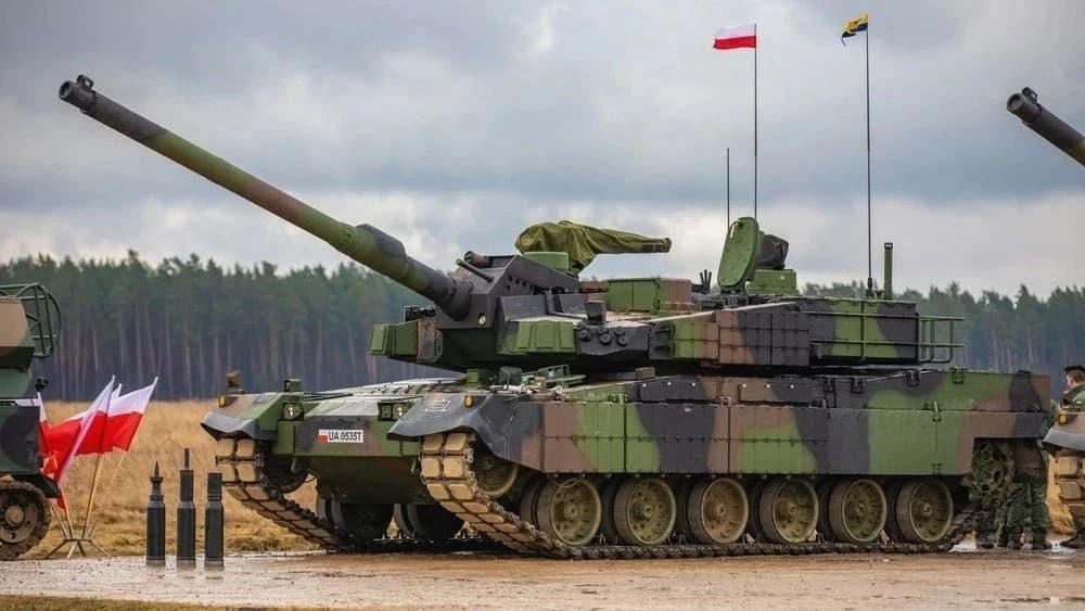 A camouflaged main battle tank flying Polish flags at a training ground